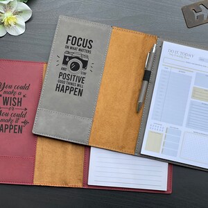 May include: A gray and brown leather-bound journal with a pen and a motivational quote on the front cover. The quote reads "Focus on what matters and stay positive, good things will happen". The journal has a daily planner section with a to-do list, a notes section, and a grid for scheduling.