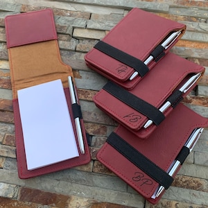 Puede incluir: Cuatro cuadernos de cuero burdeos con bandas el&aacute;sticas negras y bol&iacute;grafos plateados. Cada cuaderno tiene un nombre diferente grabado en la parte delantera: "TF", "FB", "BP".