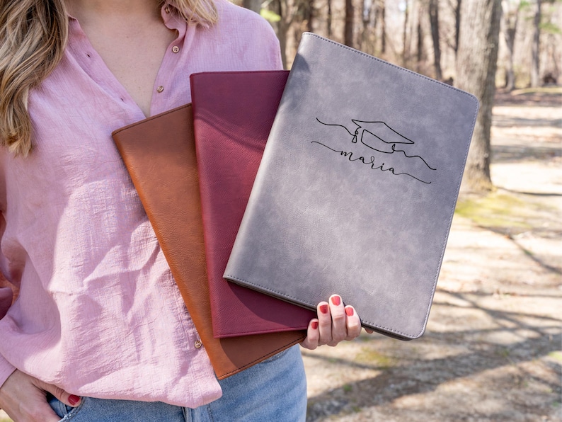 May include: A woman holds three leather-bound journals. The top journal is gray and has the name "maria" written on it with a graduation cap illustration. The middle journal is burgundy and the bottom journal is brown.