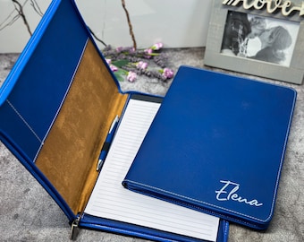 May include: A blue leather portfolio with white stitching and a zipper closure. The interior features a brown pocket and a notepad with a pen. The name "Elena" is written in white script on the front.