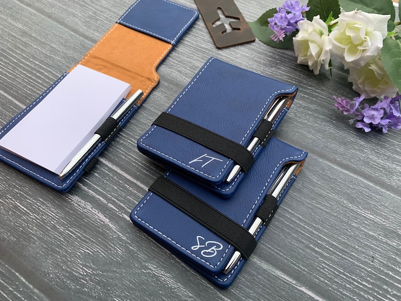 Puede incluir: Tres cuadernos de cuero azul con cierres el&aacute;sticos y bol&iacute;grafos plateados. Los cuadernos tienen costuras blancas e iniciales personalizadas.