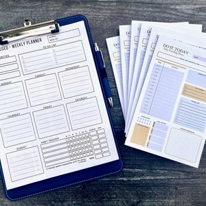 May include: A weekly planner with a blue cover and a habit tracker. The planner has a section for top priorities, a to-do list, and a daily schedule. The planner is next to a stack of daily planners with the text "DO IT TODAY" on the cover.