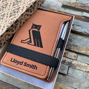Puede incluir: Un diario de cuero marr&oacute;n con un dise&ntilde;o de b&uacute;ho negro y un cierre de banda el&aacute;stica negra. El diario est&aacute; personalizado con el nombre "Lloyd Smith". Viene con un bol&iacute;grafo plateado y est&aacute; empaquetado en una caja marr&oacute;n con un patr&oacute;n floral.