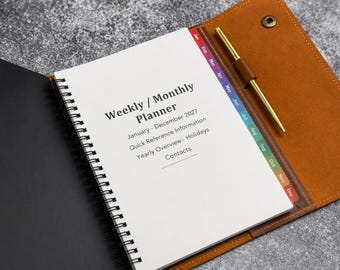 May include: A spiral-bound weekly and monthly planner for January through December 2027, featuring a tan leather cover with a snap closure, a gold pen in a loop, and colorful monthly index tabs. The white pages include sections for holidays and contacts.