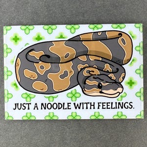 May include: A rectangular sticker featuring a cartoon snake with a brown and gray pattern. The snake is coiled on a light green background with clover-like designs. The text "JUST A NOODLE WITH FEELINGS" is below the snake.