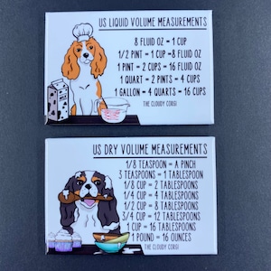 May include: Two white magnetic signs with a cartoon dog wearing a chef's hat. The signs feature US Liquid Volume Measurements and US Dry Volume Measurements with conversions for cups, pints, quarts, gallons, teaspoons, tablespoons, and ounces. The signs are decorated with cartoon images of baking supplies.