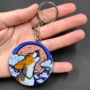 K&ouml;nnte beinhalten: Ein runder, blauer und wei&szlig;er Emaille-Schl&uuml;sselanh&auml;nger mit einem Cartoon-Beagle-Hund, der zu einem rosafarbenen Mond mit wei&szlig;en Wolken heult. Der Schl&uuml;sselanh&auml;nger hat einen silbernen Metallring und eine Kette. Der Text "The Crafty Coop" ist auf der Unterseite des Schl&uuml;sselanh&auml;ngers aufgedruckt.