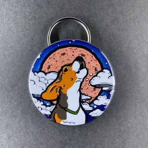 K&ouml;nnte beinhalten: Ein runder, metallener Flaschen&ouml;ffner mit einem farbenfrohen Design. Das Design zeigt einen Cartoon-Hund, der zu einem gro&szlig;en, rosafarbenen Mond mit einer keksartigen Textur heult. Der Hund ist braun, wei&szlig; und orange mit einem gr&uuml;nen Halsband. Der Hintergrund ist blau mit wei&szlig;en Wolken. Der Text "The Crafty Corgi" ist unten aufgedruckt.