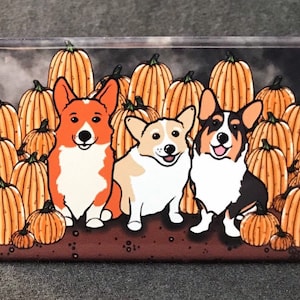 May include: Three cartoon corgi dogs wearing different colored sweaters stand in front of a pile of orange pumpkins. The corgis are smiling and looking at the viewer.