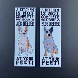 May include: Two white rectangular magnets with black text and illustrations of Australian Cattle Dogs. The text reads "A kitchen is not complete without a Red Heeler" and "A kitchen is not complete without a Blue Heeler". Both magnets have the text "At your feet!" at the bottom.