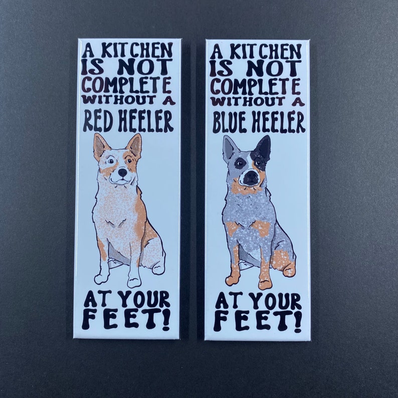 May include: Two white rectangular magnets with black text and illustrations of Australian Cattle Dogs. The text reads "A kitchen is not complete without a Red Heeler" and "A kitchen is not complete without a Blue Heeler". Both magnets have the text "At your feet!" at the bottom.