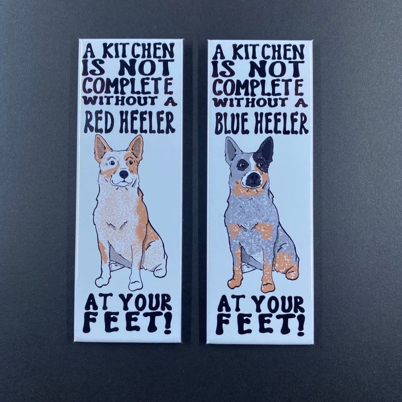 May include: Two white rectangular magnets with black text and illustrations of Australian Cattle Dogs. The text reads "A kitchen is not complete without a Red Heeler" and "A kitchen is not complete without a Blue Heeler". Both magnets have the text "At your feet!" at the bottom.