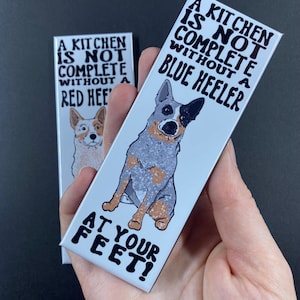 May include: Two white rectangular signs with black text and illustrations of blue heeler dogs. The top sign reads "A kitchen is not complete without a blue heeler". The bottom sign reads "at your feet!"