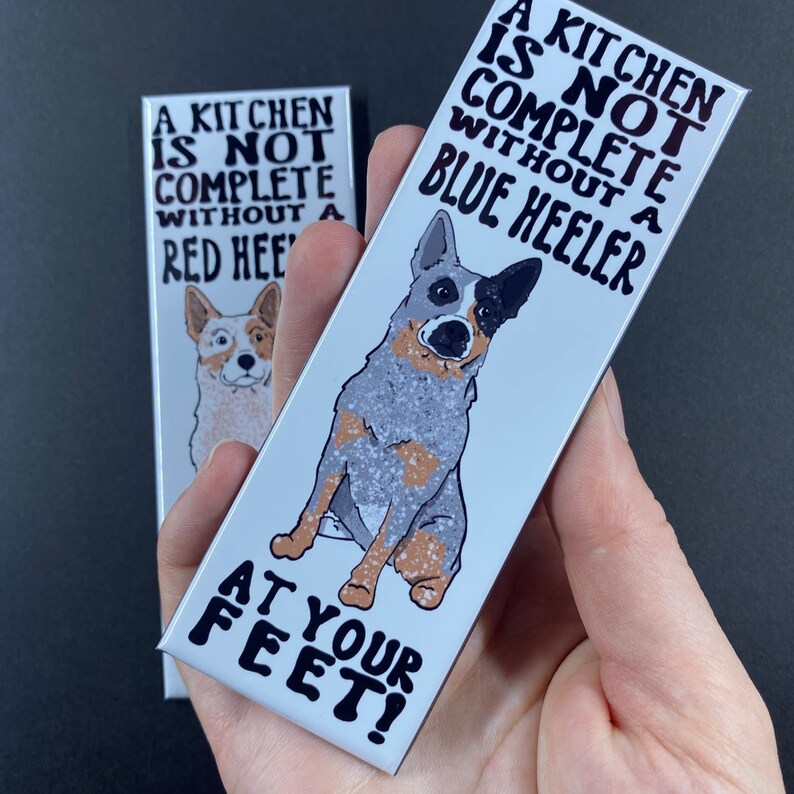 May include: Two white rectangular signs with black text and illustrations of blue heeler dogs. The top sign reads "A kitchen is not complete without a blue heeler". The bottom sign reads "at your feet!"
