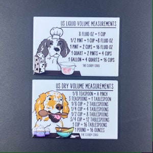 May include: Two white magnets with illustrations of dogs. The top magnet has a white and brown dog wearing a chef's hat and holding a measuring cup. The text on the magnet reads "US Liquid Volume Measurements" and includes conversions for fluid ounces, pints, quarts, and gallons to cups. The bottom magnet has a white and orange dog holding a wooden spoon and a bowl of cupcakes. The text on the magnet reads "US Dry Volume Measurements" and includes conversions for teaspoons, tablespoons, cups, and pounds to ounces.
