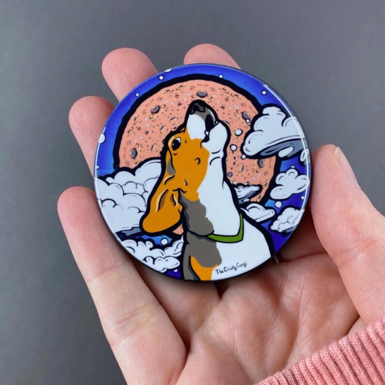K&ouml;nnte beinhalten: Ein runder, bunter Magnet mit einem Cartoon-Beagle-Hund, der zum Mond heult. Der Mond ist rosa und orange mit wei&szlig;en Wolken, die ihn umgeben. Der Text "The Cloudy Gang" ist am unteren Rand des Magneten sichtbar.