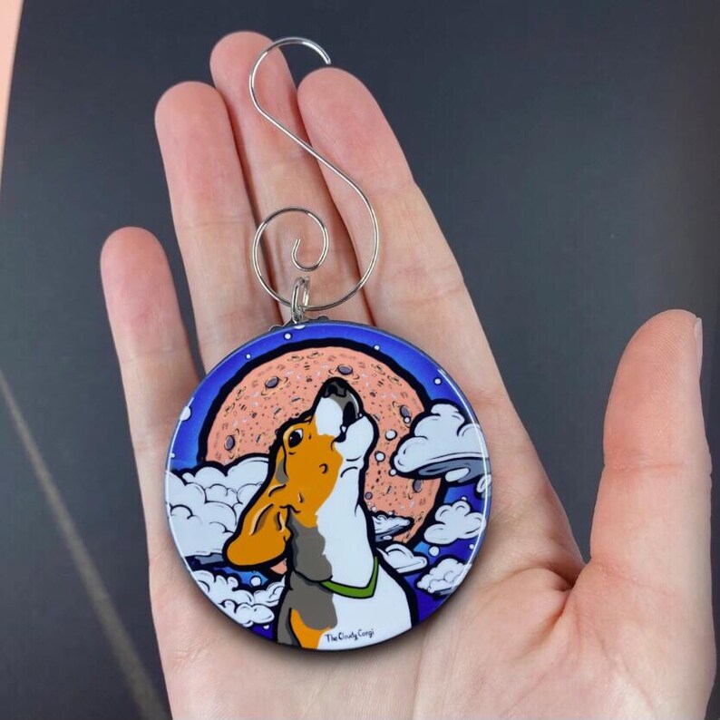 K&ouml;nnte beinhalten: Ein runder, bunter Anh&auml;nger mit einem Cartoon-Beagle-Hund, der zum Mond heult. Der Mond ist pfirsichfarben mit blauem Himmel und wei&szlig;en Wolken im Hintergrund. Der Anh&auml;nger hat einen silbernen Aufh&auml;nger.