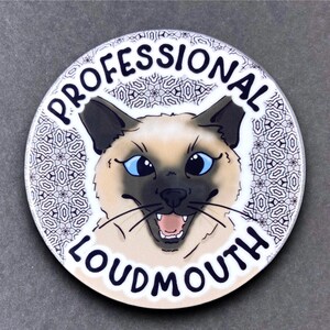 May include: A round white coaster with a black and white patterned background. A cartoon Siamese cat with blue eyes and an open mouth is in the center of the coaster. The text "PROFESSIONAL LOUDMOUTH" is written in black around the cat.