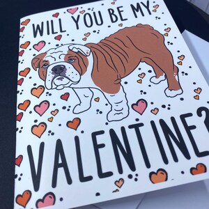 May include: A Valentine's Day card featuring a cartoon bulldog with the text "Will you be my Valentine?" surrounded by pink, orange, and yellow hearts.