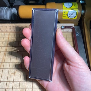 May include: A rectangular black magnet with a silver frame. The magnet is being held in a hand.