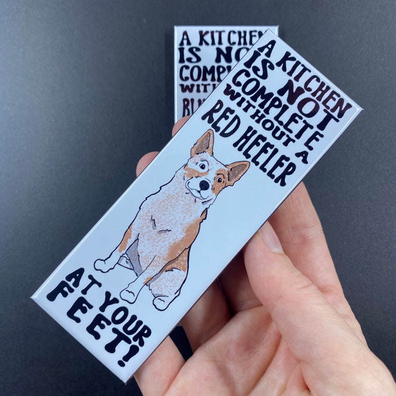 May include: A white rectangular magnet with a cartoon red heeler dog and the text "A kitchen is not complete without a red heeler at your feet!"
