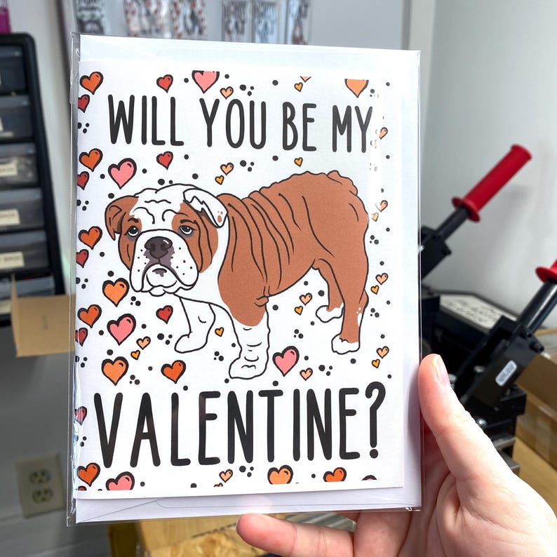 May include: A white card with a brown and white bulldog illustration and the text "Will you be my Valentine?" surrounded by pink and orange hearts.