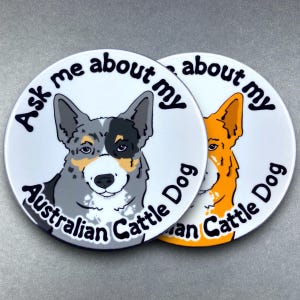 May include: Two round white coasters with black text and a cartoon dog on each. The text on the left coaster reads "Ask me about my Australian Cattle Dog". The text on the right coaster reads "Ask about my Australian Cattle Dog".