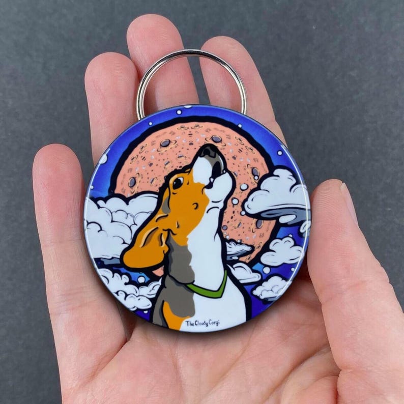 K&ouml;nnte beinhalten: Ein runder, blau-wei&szlig;er Schl&uuml;sselanh&auml;nger mit einer Cartoon-Illustration eines Hundes, der zum Mond heult. Der Mond ist pfirsichfarben mit einer gesprenkelten Oberfl&auml;che. Der Text "The Cloudy Corp" ist auf der Unterseite des Schl&uuml;sselanh&auml;ngers aufgedruckt.