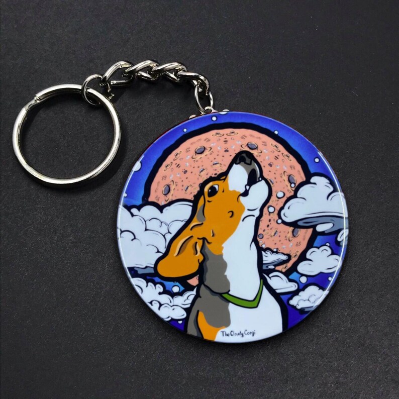 K&ouml;nnte beinhalten: Ein runder, blau-wei&szlig;er Schl&uuml;sselanh&auml;nger mit einem Cartoon-Beagle, der zum Mond heult. Der Mond ist rosa und orange mit einer wei&szlig;en Wolke davor. Der Text "The Crazy Corgi" befindet sich am unteren Rand des Schl&uuml;sselanh&auml;ngers.