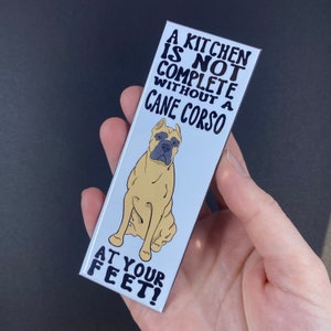 May include: A white rectangular magnet with black text that reads "A kitchen is not complete without a Cane Corso at your feet!" and a cartoon image of a tan Cane Corso dog with a grey nose and black eyes sitting on a white background.