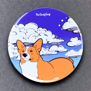 May include: A round, blue and white button with a cartoon image of a corgi dog with a white chest and orange body. The corgi is looking to the left and is surrounded by white clouds on a blue background. The text "The Cloudy Corgi" is at the top of the button.