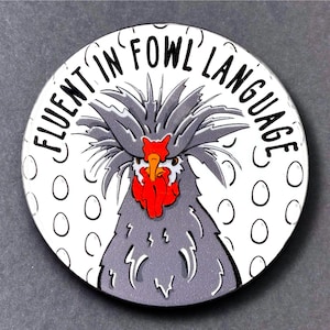 May include: A white round button with a gray and red rooster illustration and the text "Fluent in Fowl Language".