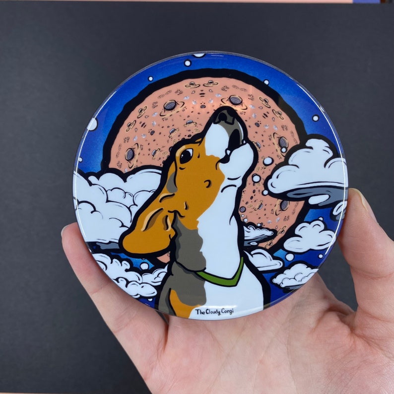 K&ouml;nnte beinhalten: Ein runder, blauer und wei&szlig;er Untersetzer mit einem Cartoon-Beagle, der zu einem rosa-braunen Mond heult. Der Mond hat eine gesprenkelte Oberfl&auml;che und der Hund tr&auml;gt ein gr&uuml;nes Halsband. Der Untersetzer tr&auml;gt den Text "The Cloudy Corgi" am unteren Rand.