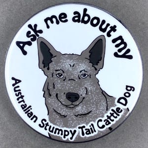May include: A round, white button with a cartoon illustration of an Australian Stumpy Tail Cattle Dog. The text "Ask me about my Australian Stumpy Tail Cattle Dog" is printed around the dog's image in black.