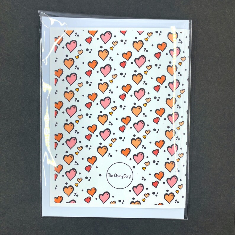 May include: A white card with a repeating pattern of pink and orange hearts on a light blue background. The card has the text "The Cloudy Corgi" in a circle at the bottom.