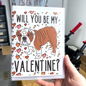 May include: A white card with a brown and white bulldog illustration. The card has a pink and orange heart pattern and the text "Will you be my Valentine?"