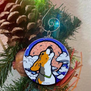 K&ouml;nnte beinhalten: Ein runder Weihnachtsbaumschmuck mit einem Cartoon-Hund, der zum Mond heult. Der Hund ist braun, wei&szlig; und schwarz mit einem gr&uuml;nen Halsband. Der Mond ist rosa mit braunen Flecken und von wei&szlig;en Wolken umgeben. Der Schmuck hat einen blauen Hintergrund mit wei&szlig;en Schneeflocken. Der Text "The Crafty Corgi" ist auf der Unterseite des Schmucks aufgedruckt.