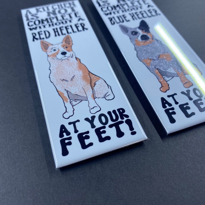 May include: Two white rectangular magnets with a cartoon image of a red heeler and a blue heeler dog. The text on the magnets reads "A kitchen is not complete without a red heeler" and "A kitchen is not complete without a blue heeler". Both magnets have the text "At your feet!" at the bottom.
