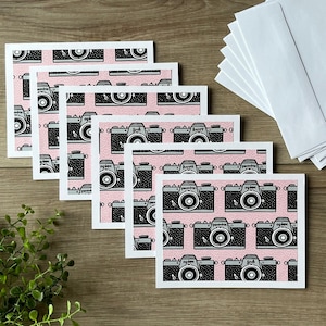 May include: A set of greeting cards with a repeating pattern of black and gray cameras on a pink background. The cards are rectangular with white borders and are accompanied by a stack of white envelopes. The word "happy" is visible on some of the cameras.
