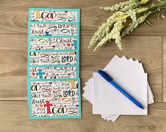 May include: A stack of greeting cards with a teal border and a patterned design featuring religious words and symbols. A blue pen and blank white cards are next to the stack. A sprig of white flowers is in the background.