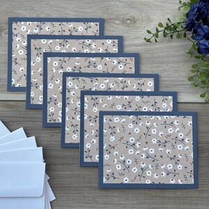 May include: Set of floral greeting cards with a blue border. The cards feature a beige background with a delicate white floral pattern. A stack of white envelopes is visible in the lower left corner. The cards are arranged on a wooden surface.