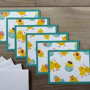 May include: Six blank greeting cards with a teal border and a pattern of yellow chicks in various poses on a white background with small white dots. The chicks are wearing sunglasses, hats, and some are holding hearts.