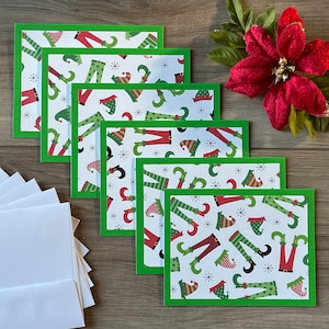 May include: A set of six Christmas greeting cards with a green border and a festive pattern of elf legs and hats on a white background. White envelopes are visible on the left, and a red poinsettia is on the right.