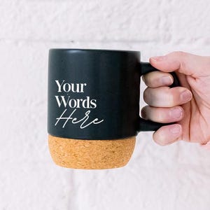 May include: Black coffee mug with a cork base, featuring the text "Your Words Here" in white script. The mug is held by a person wearing a colorful sweater against a white brick wall.