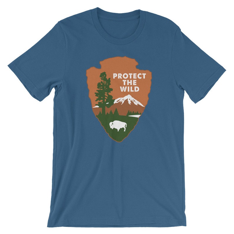 May include: A blue t-shirt with a brown and green National Park Service arrowhead logo. The logo features a mountain, a tree, a lake, and a buffalo. The text "PROTECT THE WILD" is printed in white on the logo.
