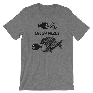 May include: A heather grey t-shirt with a black graphic of two fish. The smaller fish is being swallowed by the larger fish, which is made up of many smaller fish. The text "ORGANIZE!" is printed below the graphic.