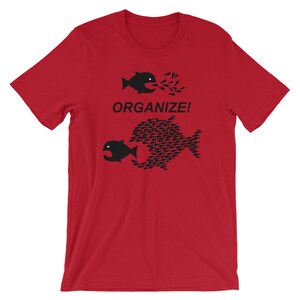 May include: Red t-shirt with a black graphic of two fish. The smaller fish is being swallowed by the larger fish, which is made up of many smaller fish. The text "ORGANIZE!" is printed below the fish.