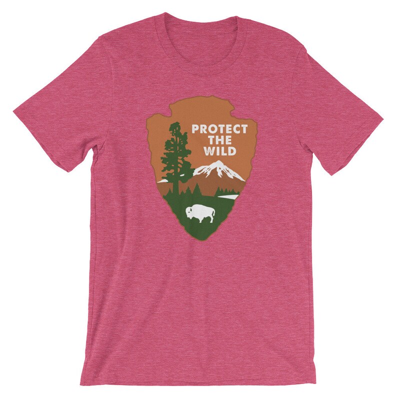 May include: A pink heather t-shirt with a green, brown, and white graphic of the National Park Service arrowhead logo. The logo features a mountain, a tree, and a buffalo. The text "PROTECT THE WILD" is printed in white on the logo.