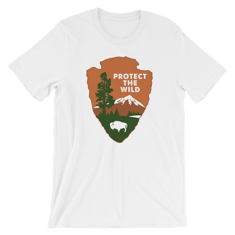 May include: White t-shirt with a brown and green graphic of the National Park Service arrowhead logo. The logo features a mountain, a tree, a lake, and a buffalo. The text "PROTECT THE WILD" is printed in white on the logo.
