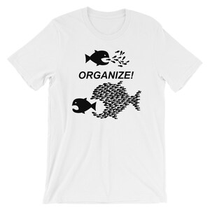 May include: White t-shirt with a black graphic of two fish. The top fish is small and being eaten by a larger fish made up of many smaller fish. The text "ORGANIZE!" is printed below the fish.
